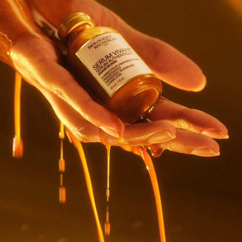 Hand holding bottle of Biologique Recherche Serum Vivant, an amber-liquid. On the bottle is a white label with a gold border, featuring text and the Biologique Recherche logo. Freeflowing amber liquid pours down from the hand.